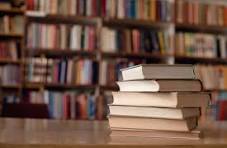 A stack of books resting on a table in front of a library shelf